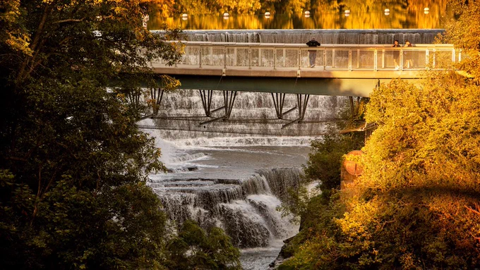 Sunlight and trees surround the falls and foot bridge at Beebe Lake at Cornell University.