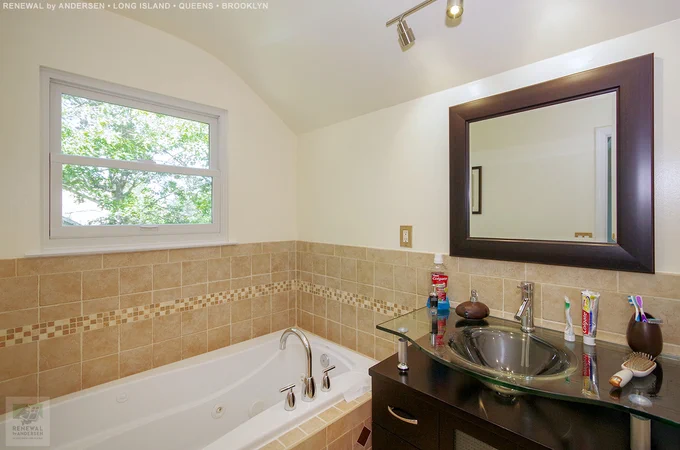 Attractive bathroom with new double hung window we installed. This rich and handsome bathroom with dark wood cabinets and framed mirror, looks wonderful with this new white window we installed, against cream color walls, beige tiles and white fixtures. Find out more about getting new windows from Renewal by Andersen of Long Island, serving Suffolk, Nassau, Queens and Brooklyn.