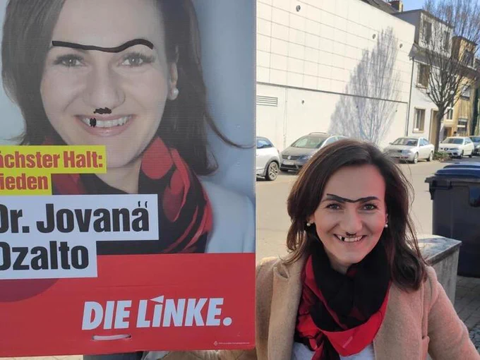 Linke Jovana Dzaltos Wahlplakat wurde mit einer Monobraue und einem schwarzen Zahn bemalt. Sie hat dies auf ihr Gesicht abgemalt und posiert neben dem Plakat.