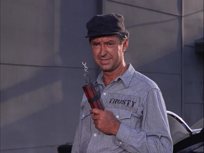 a man named trustee with his name written on his great jumpsuit, hold on to a stick of dynamite.