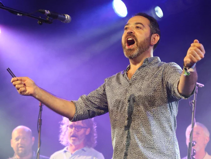 Paul Sartin singing on stage with a microphone in front of him. You can see a few other people in the background singing behind him.