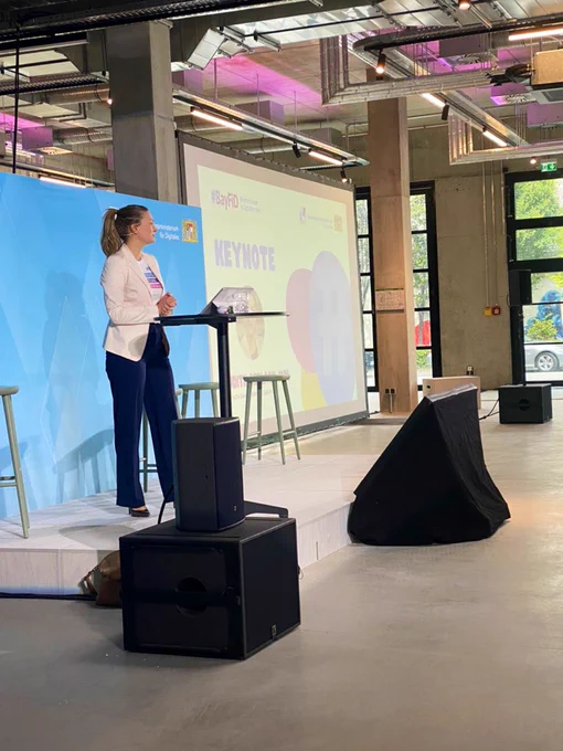 Eine Frau (die bayerische Digitalministerin Judith Gerlach) in dunkler Hose und weissem Blazer steht auf einer Bühne. Im Hintergrund eine Leinwand in weiss und blau auf der „#BayFiD“ und „Keynote“ steht.