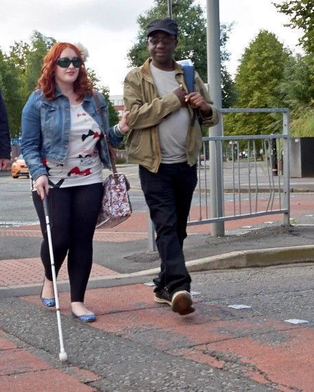 Image shows a woman crossing a pedestrian crossing, holding onto a man's elbow for guiding and holding a long cane.
