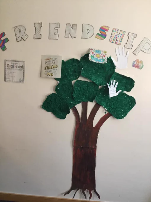 A tree made from painted paper on a wall, with colourful letters above spelling out 'Friendship' and pieces of paper with messages stuck around it.