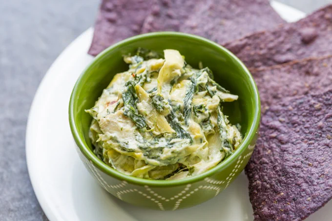 Hot spinach artichoke dip in a green bowl on a white plate with an array of blue corn tortilla chips