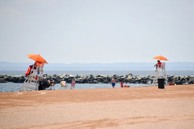 Lifeguards look out to folks on the beach and in the water from their lifeguard chairs as beachgoers enjoy walking along the shores and swimming in the water.