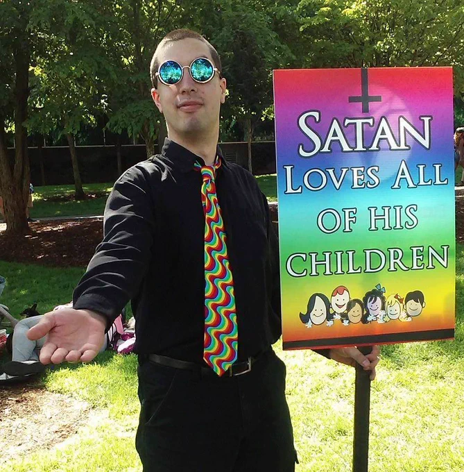 Man in sunglasses and tie holding a sign that says "Satan Loves All Of His Children"