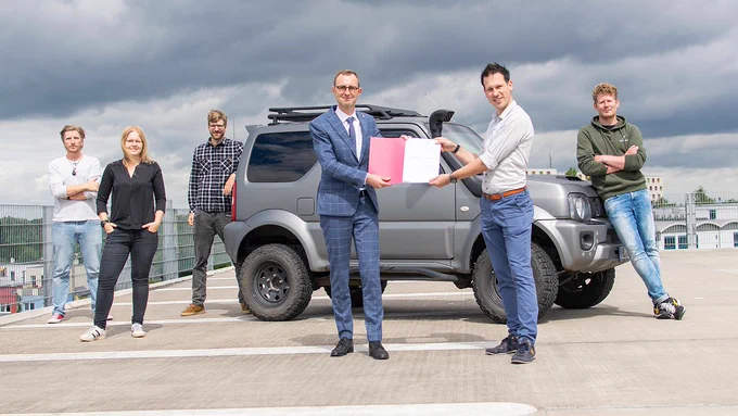 Staatssekretär Dirk Schrödter überreicht einen Förderbescheid an Sascha Klement. Im Hintergrund stehen mehrere Mitarbeiter der Firma um einen Geländewagen herum. 