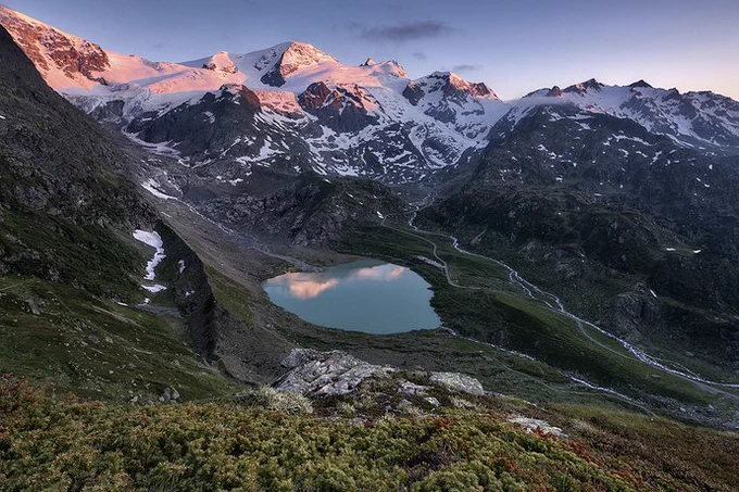 Photo of the Alps mountains with a lake and sunlight setting on the distant mountains
