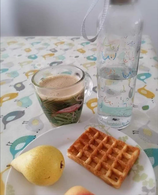 A clear water bottle with coloured boob outlines, a glass cup of cappuccino next to a plate with a waffle, apricot and pear, on a table covered in a simple bird print tablecloth. 