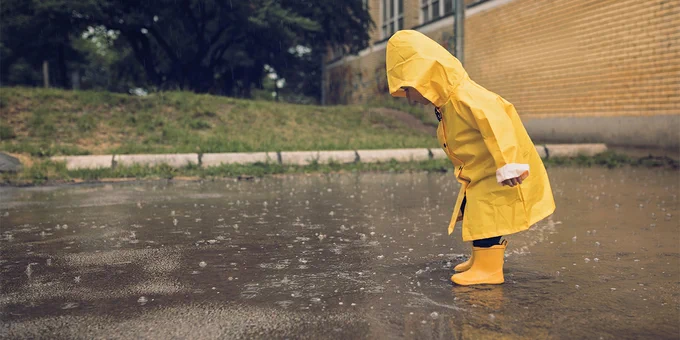 Afbeelding: Een kind in een geel regenjas en gele laarzen die in de regen in een plas aan het staan is.