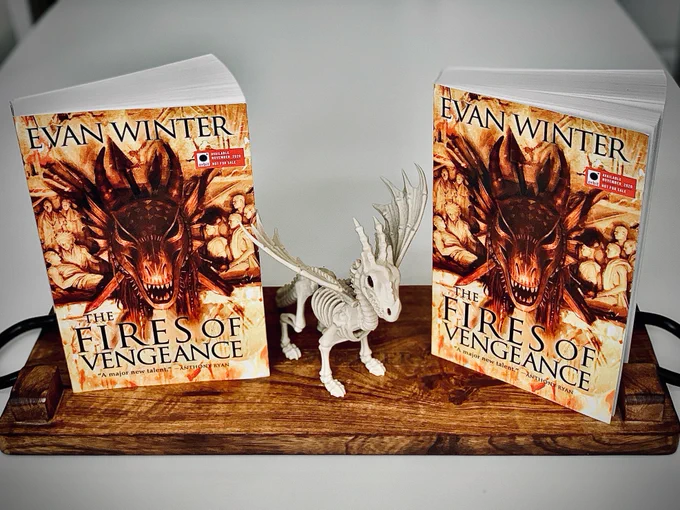 Two copies of THE FIRES OF VENGEANCE paperback standing up on a wooden board. Between the two books there is a dragon skeleton (presumably plastic and not real).