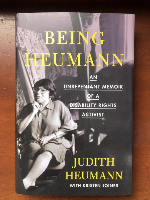 Book cover: text is Being Heumann. An unrepentant memoir of a disability rights activist. Judith Heumann. With Kristen Joiner. Image of Judith wearing glasses and sitting in a wheelchair looking directly at camera. This image is on left hand side of cover and is highlighted on other images which are greyed our and include a silhouette of a person and curtains.