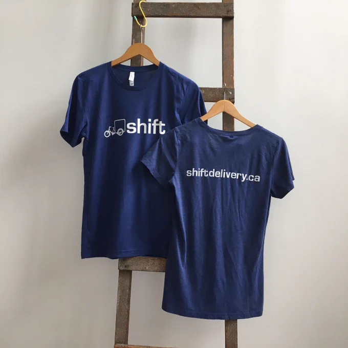 Two blue Shift Delivery t-shirts displayed hanging from a ladder in front of a white wall.