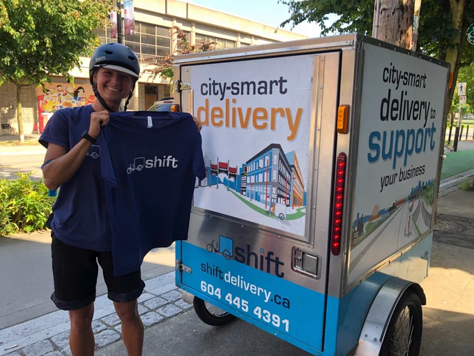 Shift Delivery worker holding up blue t-shirt with Shift logo, standing outside of VALU CO-OP's Chinatown Studio and in front of their bike cargo trailer.