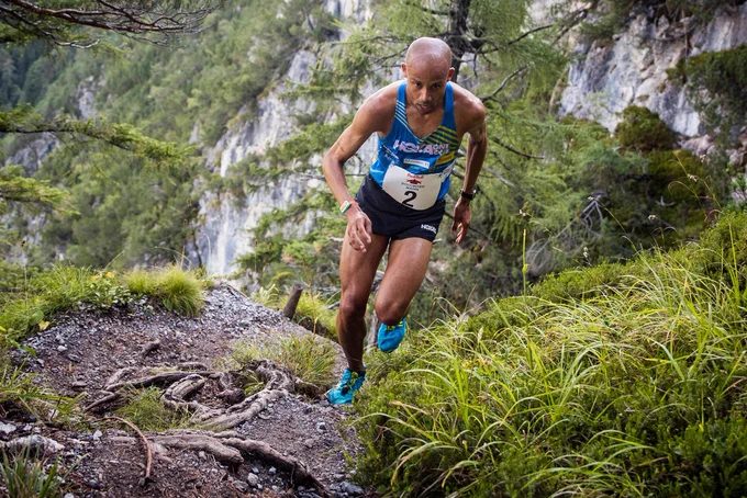 Joe racing on steep and rugged terrain. He is running over rocks and roots along a mountain trail. His focus is straight ahead, his arms and knees bent, propelling him forward with power in each stride. He is wearing a blue HOKA ONE ONE singlet, black shorts, blue shoes, and a white racing big with the number 2 on it.