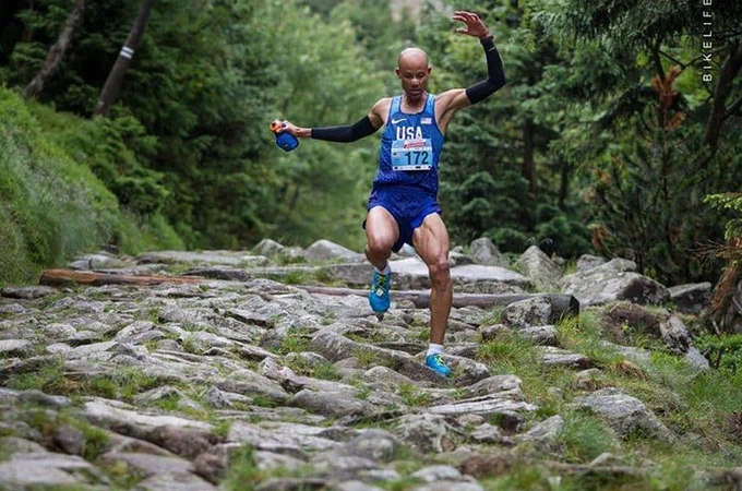 Joe flying down a rocky descent in the mountains. His arms are out wide, to his side and over his head. He is wearing a blue Team USA singlet and shorts and blue shoes, with black compression sleeves and a water bottle in his right hand. Picture credit: Bike Life.