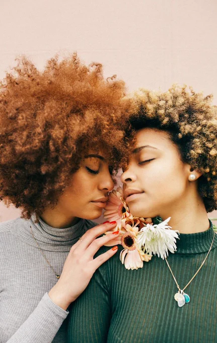 Imagem de duas mulheres negras apaixonadas, elas estão com os rostos próximos e seguram flores nas mãos, seus olhos estão fechados e seus cabelos crespos estão soltos.