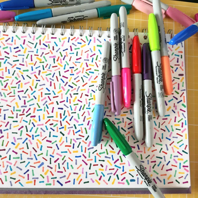 Coloured Sharpie pens on top of a sketchbook with a hand drawn pattern that looks like sprinkles