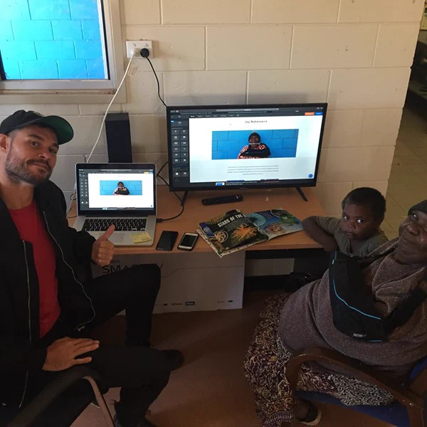 Greg, an inDigiMOB Digital Access Worker, and Joy, an Aboriginal artist, sitting with a computer on a desk in between them that is showing Joy's new website on the screen, which was created to promote her art.