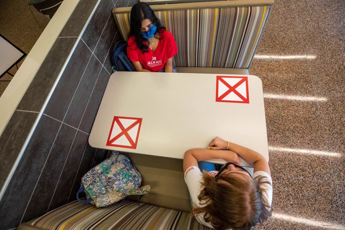 Overhead view of two students at a booth in Martin Dining. Students are sitting diagonally from each other, with large X decals on the empty spaces. 