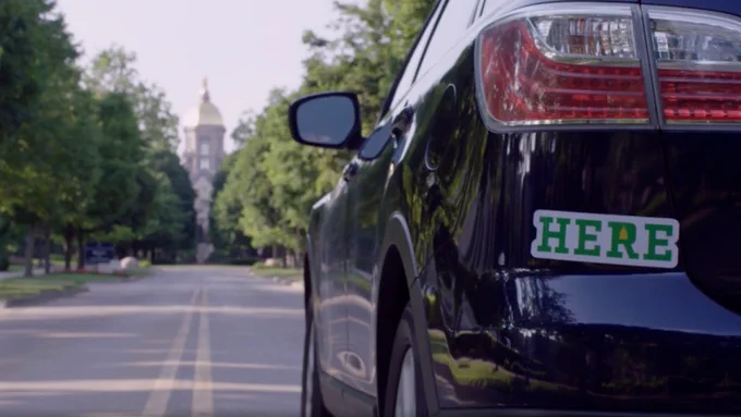 A black car with a bumper sticker reading "Here" driving towards the Golden Dome of the University of Notre Dame
