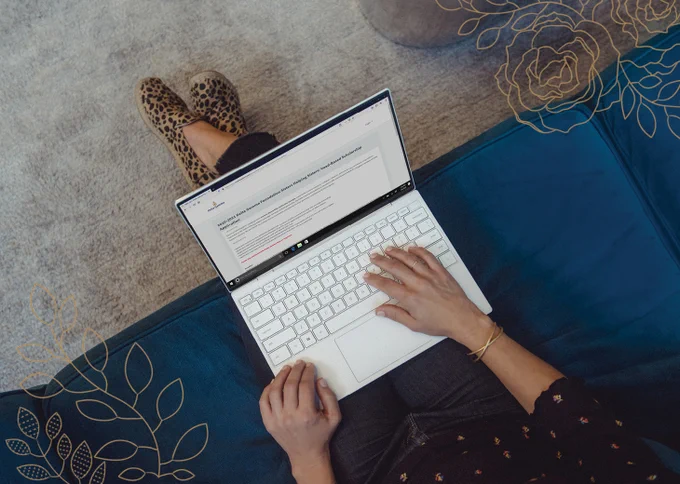 Overhead photo of a woman sitting on a couch with a laptop on her lap. The browser is open to the Sisters Helping Sisters application page.