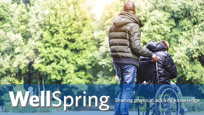 A man is pushing another adult in a wheelchair on a shared path in the spring