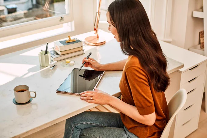 A woman is sitting at her desk in her bright sun-filled apartment using her Lenovo Yoga like a tablet.