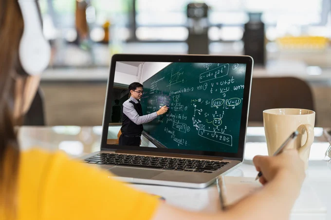 View of student watching professor teach physics online, writing equations on a blackboard.