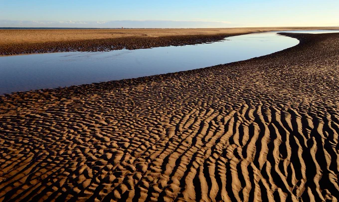 Looking across a beach with wavy sand and a small river of water going towards the sea