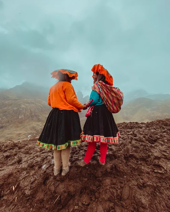 Two people wearing black skirts and vibrant shirts are shown from behind, standing on top of a hill. Cloudy skies cover the mountains ahead of them. The two people are holding hands. One smiles and laughs while looking at the other.