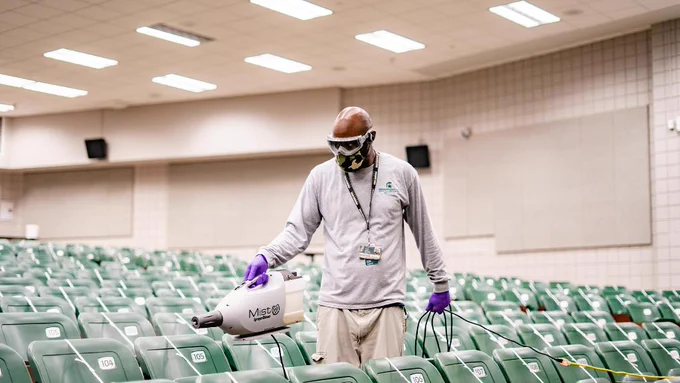 An IPF employee wearing safety goggles, a mask and gloves sanitizing seats in a classroom