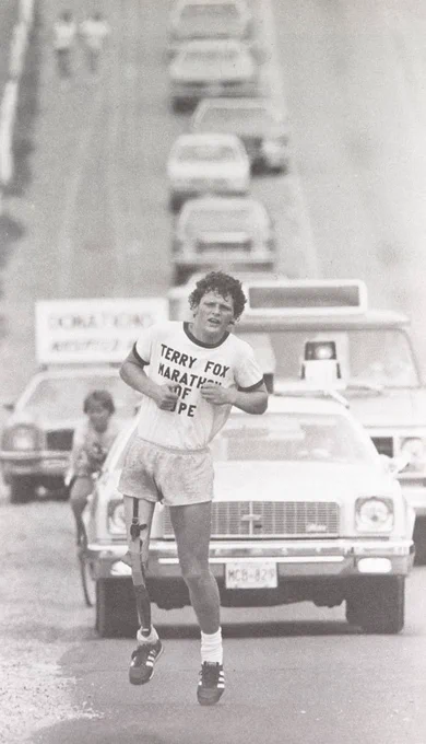 Black and white photo of Terry Fox running down a road as cars follow him from behind 
