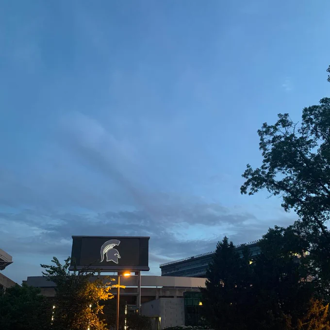 A photo of Spartan Stadium at sunset of dark blue skies and clouds up top, the Spartan helmet logo glowing and tree branches on the right side