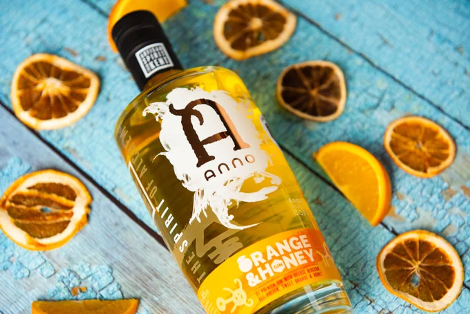 A bottle of Anno Orange & Honey Gin, laying on a blue painted, old wooden table, surrounded by slices of fresh and dried orange segments.