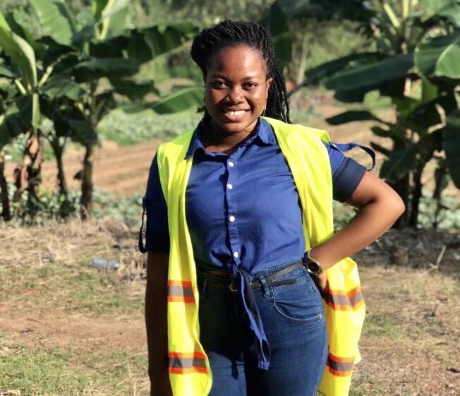 A smiling picture of Recca Ebe wearing a fluorescent jacket, dark blue shirt and jeans She is standing in a field with plantain plants in the background