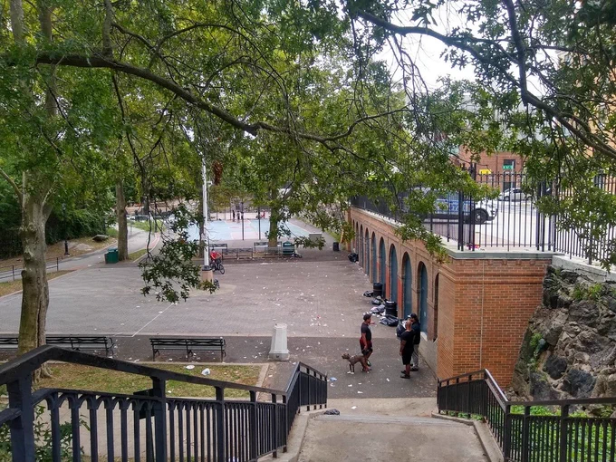 Looking down the stairs to the park, which has trash on the ground. An NYPD SUV can be seen on the sidewalk to the right, above and just outside of the park.