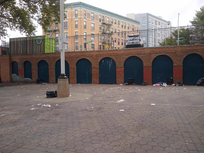 Looking at the trash on the ground at the park. An NYPD SUV can be seen on the sidewalk above and just outside of the park.