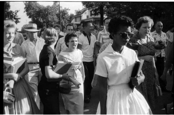 Elizabeth Eckford (in a white dress, carrying a school folder and wearing sunglasses) walking past Central High School with a Central student named Hazel Bryan (directly behind Elizabeth, with clenched teeth and wearing a white dress) yelling at her in the midst of an angry mob.

