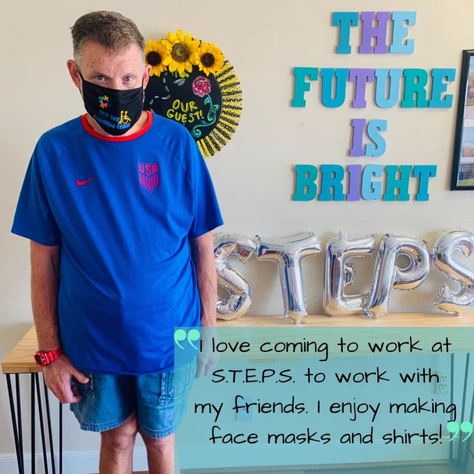 Our new employee of the month, wearing a black mask, blue shirt, and blue jean shorts, standing in front of a sign that says The Future is Bright and a yellow and black wreath.