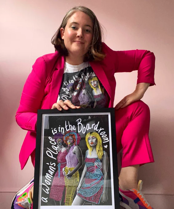 A woman (Jennifer Hayashi) wearing a bright pink business suit and neon trainers with a T-shirt that says “ a woman’s place is in the Boardroom” which a matching print in a frame in from of her looking pretty bad ass and strong!