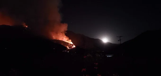 The moon rises over the mountains as flames from the fire create a night time glow.