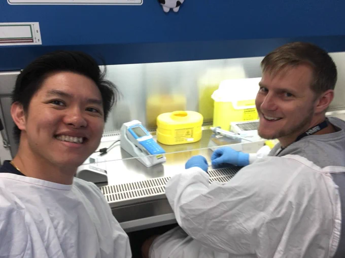 one man of asian appearance and one man of white appearance, both wearing lap coats and working with samples in a fume hood in a science lab