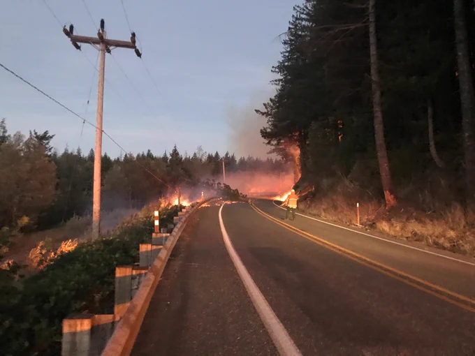 A picture of SR 410 east of Enumclaw during the dawn hours and a fire burning on both sides of the road. One person in a safety vest and hard hat sets up traffic control.