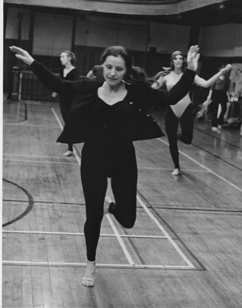 Image description: A black and white photo of a young Claire Mallardi teaching a dance class in the former Radcliffe gym. She has been captured in mid-movement on one leg with the other leg bent at the knee about to take the next step. Both arms are raised to her side and slightly bent at the elbow like wings; her gaze is slightly down towards the floor and she is facing the camera with students following her example behind her. She is wearing tights and a leotard with an open cardigan sweater over the leotard; she is barefoot and her brunette hair is tied loosely back in a pony tail.