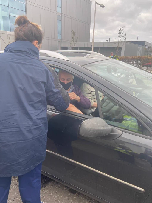 A man wearing a face mask getting a flu jab through the window of a car from a nurse.