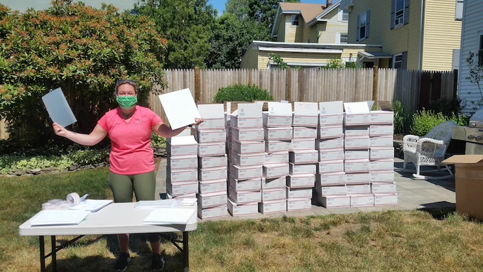 A woman in a Covid-19 masks stands in front of a large wall made of stacks of USPS priority shipping boxes