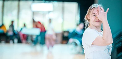 A photo of Hannah Sampson, our dancer with Down's Syndrome looking into the camera. She is white, has blond hair, and soft eyes smiling slightly. She is brushing her fringe hair off of her forehead with her left hand. She is wearing a white t-shirt