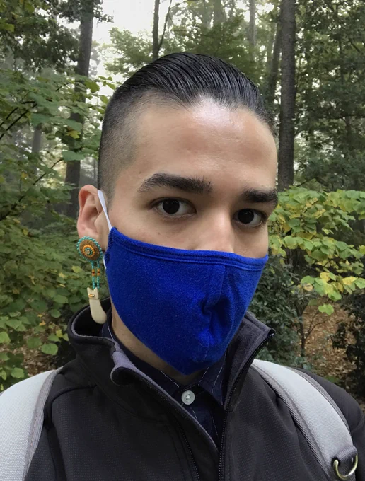 Selfie of person outside wearing a cloth mask, and wearing round earrings with elk tooth shaped dangles. Background is forest trees.